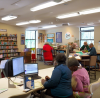 A cozy public library setting where diverse individuals, including elderly people and young adults from various ethnic backgrounds, sit at tables receiving assistance from friendly, professional tax advisors. The room is equipped with computers, tax forms, and informational posters on the walls explaining free tax services. There is a visible sign in the forefront saying, Free Income Tax Help Here! Bright and welcoming atmosphere.